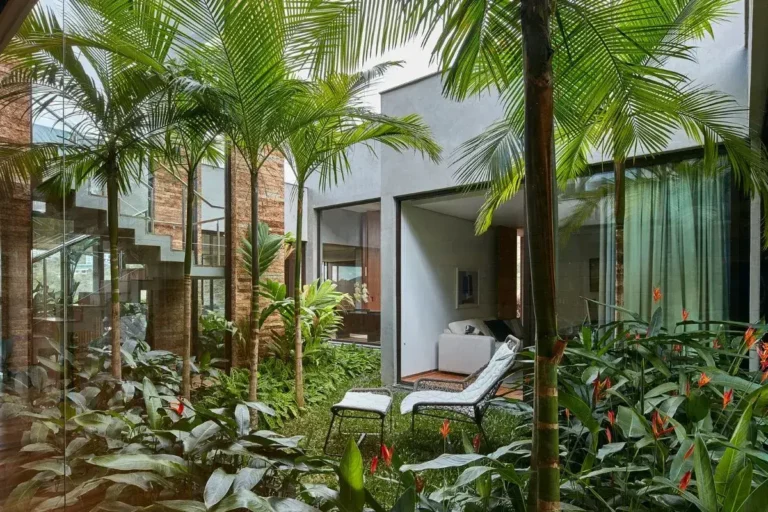 jardim com muitas espécies de plantas tropicais rodeando uma cadeira de descanso branca com parede da casa branca e de tijolo ao fundo.