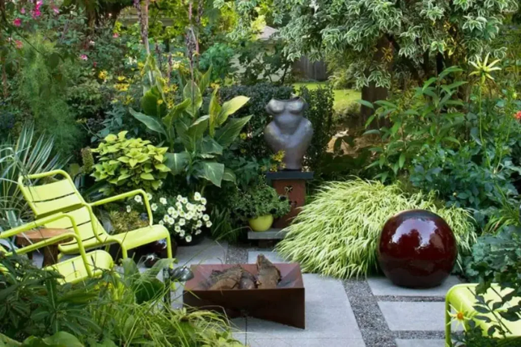 Como decorar um jardim pequeno com plantas de vários tamanhos e cores, duas cadeiras verdes, uma lareira, um busto e uma fonte em forma circular